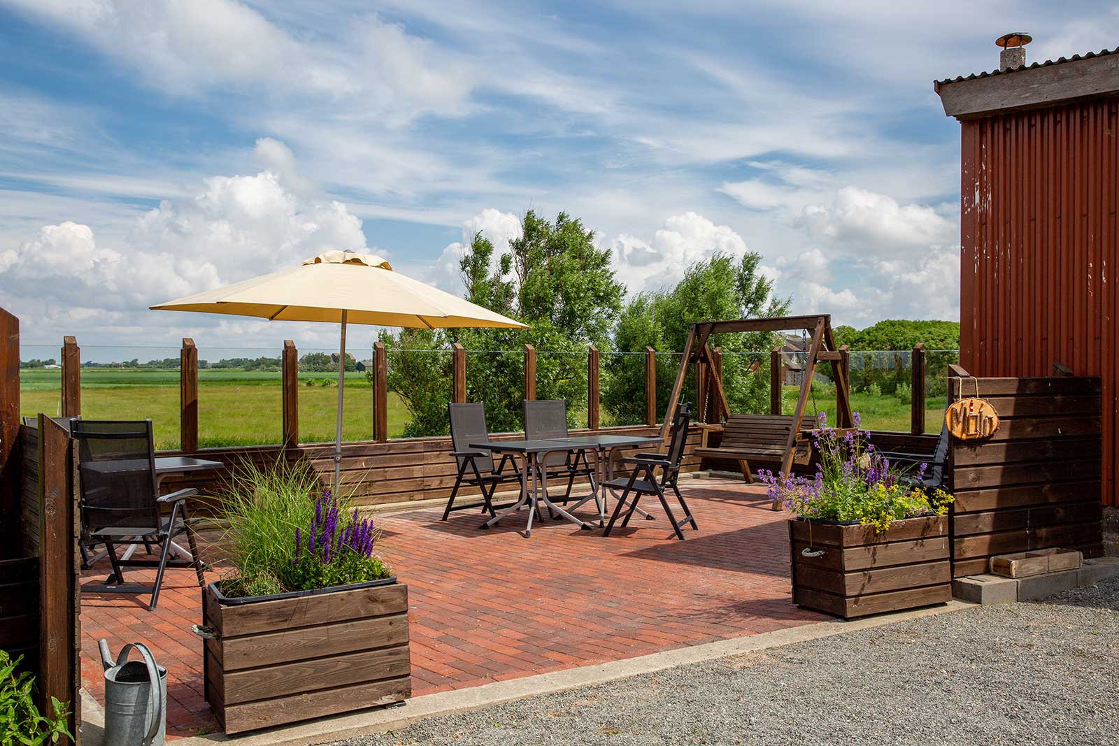 Sonnige Terrasse einer Ferienwohnung auf der Nordseeinsel Pellworm mit gelbem Sonnenschirm, Sitzgruppe, Hollywoodschaukel und blühendem Lavendel, dahinter der weite Blick über die grüne Marschlandschaft Nordfrieslands